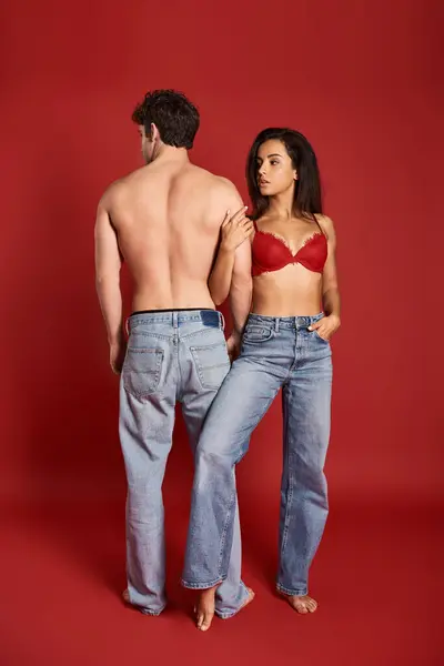 Belle jeune femme en lingerie sexy et jeans posant avec petit ami musclé sexy sur fond rouge — Photo de stock
