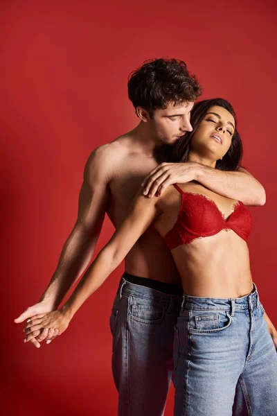 Good looking and muscular man without shirt seducing young brunette woman on red background — Stock Photo