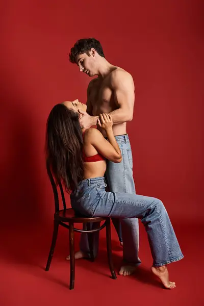 Shirtless and muscular man with closed eyes embracing brunette woman on chair on red background — Stock Photo