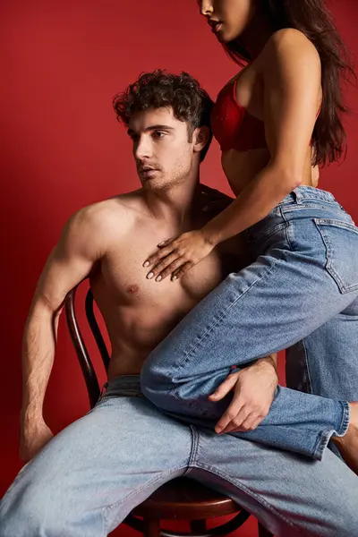 Sensual mujer joven en sujetador de encaje sentado en la parte superior de sexy novio muscular en la silla, fondo rojo - foto de stock