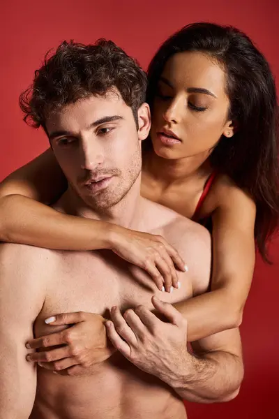 Brunette and beautiful young woman embracing her handsome muscular boyfriend on red background — Stock Photo