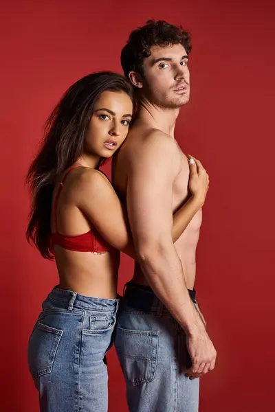 Brunette and charming young woman in lace bra and jeans embracing her muscular man on red background — Stock Photo