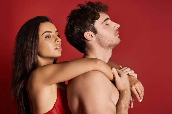 Брюнетка и очаровательная молодая женщина в кружевном лифчике обнимает своего мускулистого мужчину на красном фоне — стоковое фото