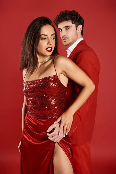 Handsome man in formal wear embracing beautiful young woman in shiny dress on red background — Stock Photo