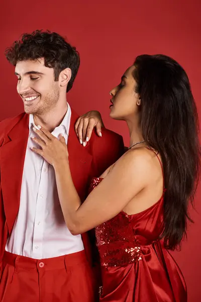 Elegante Frau mit brünetten Haaren umarmt fröhlichen Mann in formeller Kleidung auf rotem Hintergrund, Glamour — Stockfoto
