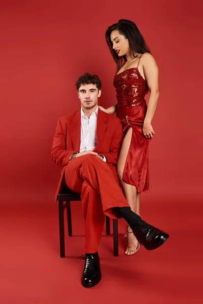 Morena mujer joven en vestido brillante de pie cerca de hombre en ropa formal en la silla, fondo rojo - foto de stock