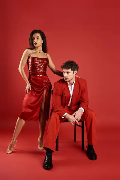 Elegante mujer joven en vestido sexy de pie cerca del hombre en el desgaste formal sobre fondo rojo, sofisticado - foto de stock