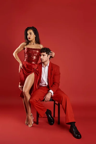 Femme élégante en robe sexy debout près de l'homme en tenue formelle sur fond rouge, sophistiqué — Photo de stock