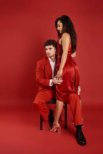 Bel homme assis sur la chaise et embrassant élégante jeune femme en robe sur fond rouge — Photo de stock
