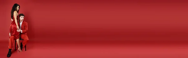 Elegant young woman in sexy dress standing near man in formal wear on red background, banner — Stock Photo