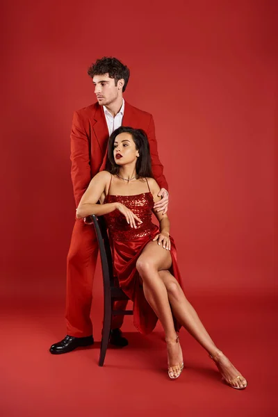 Sofisticada mujer joven en vestido sexy sentado en la silla cerca del hombre en el desgaste formal sobre fondo rojo - foto de stock