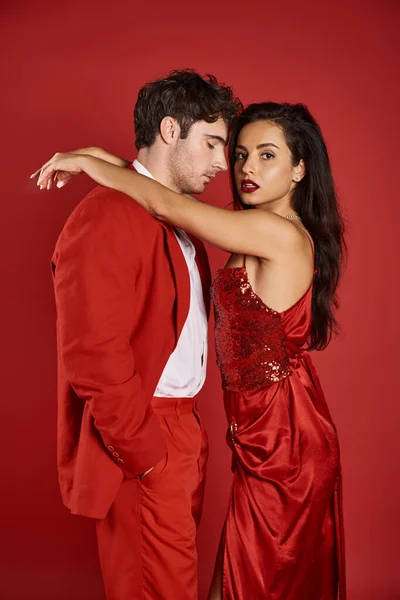 Mujer joven sofisticada y morena en vestido sexy abrazando hombre guapo sobre fondo rojo - foto de stock