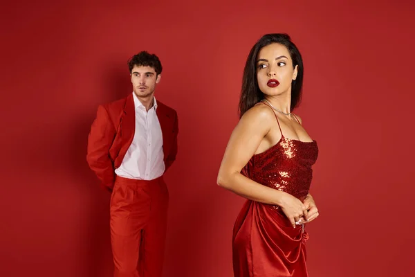 Зосередитися на чудовій і молодій жінці, позуючи в сексуальній сукні біля розмитого чоловіка в костюмі на червоному тлі — стокове фото