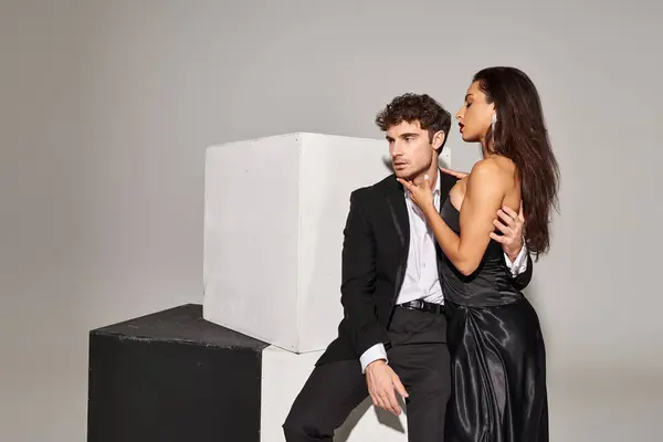 Hombre guapo en ropa formal sentado en el cubo y abrazando atractiva mujer joven en vestido de deslizamiento negro - foto de stock