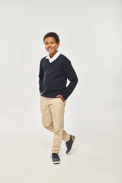 Happy african american schoolboy in neat uniform standing with hands in pockets on grey backdrop — Stock Photo