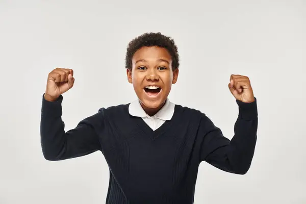 Excité afro-américain écolier en uniforme se réjouissant tout en regardant la caméra sur fond gris — Photo de stock