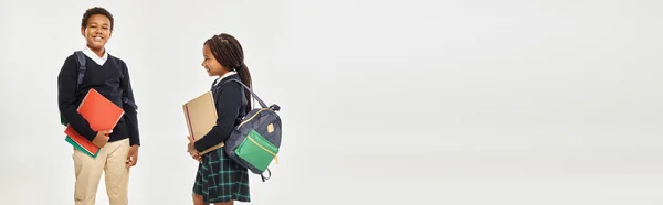 Cheerful african american kids in school uniform holding textbooks and standing on grey, banner — Stock Photo