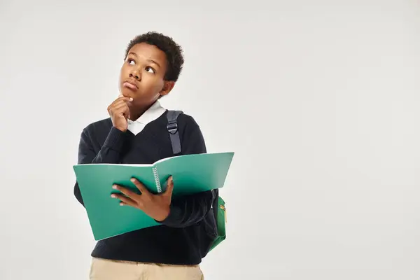 Thoughtful african american schoolboy in uniform holding notebooks and standing on grey background — Stock Photo