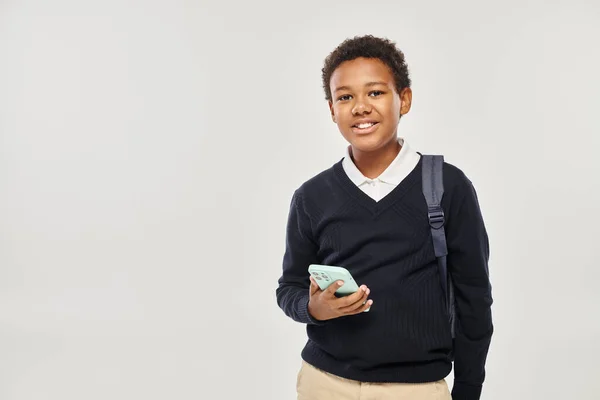 Happy african american schoolboy in uniform holding smartphone and standing on grey background — Stock Photo