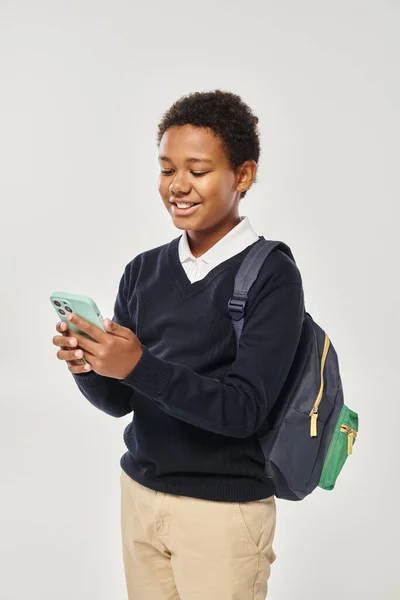 Happy african american schoolboy in uniform using smartphone and standing on grey background — Stock Photo
