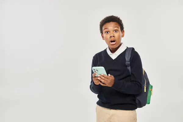 Shocked african american schoolboy in uniform holding smartphone and standing on grey background — Stock Photo