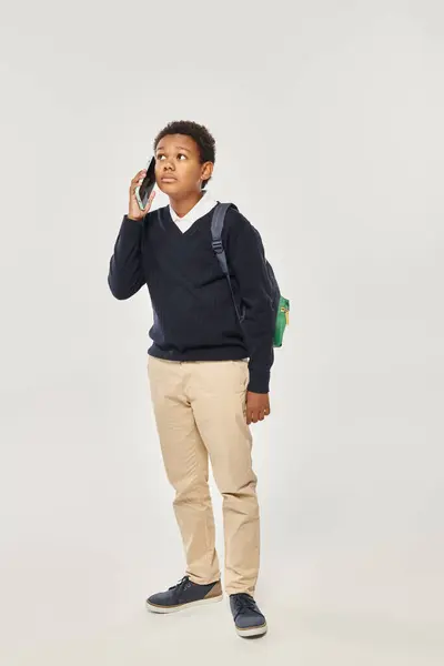 Африканский американский школьник в форме разговаривает по смартфону и стоит на сером фоне — стоковое фото