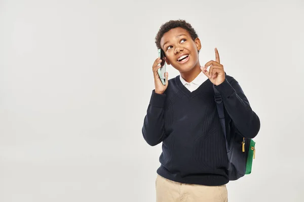 Happy african american schoolboy in uniform talking on smartphone and gesturing on grey background — Stock Photo