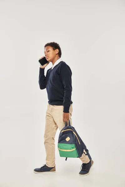 African american schoolboy in uniform talking on smartphone and walking with backpack on grey — Stock Photo
