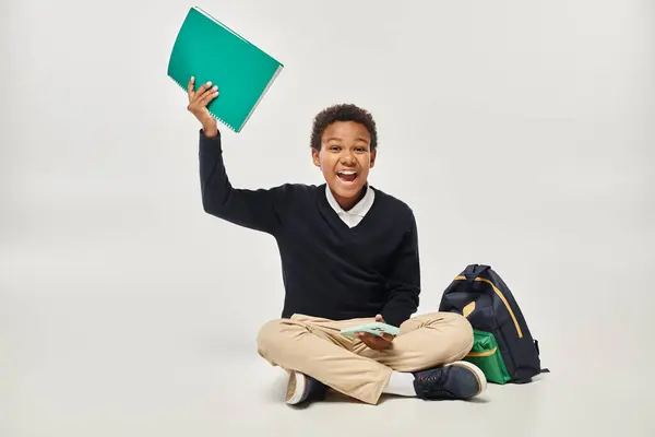 Happy african american boy in uniform holding smartphone and notebook while sitting near backpack — Stock Photo