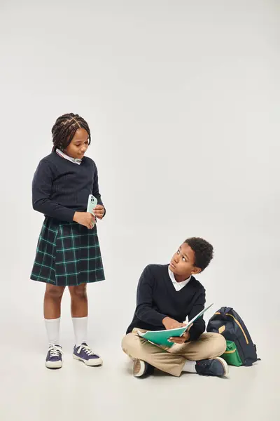 African american schoolgirl holding smartphone near schoolboy studying with notebook on grey — Stock Photo