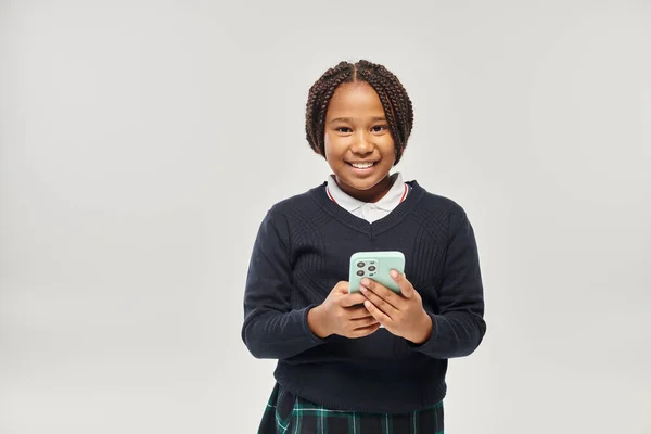Happy preteen african american schoolgirl in uniform holding smartphone on grey background — Stock Photo