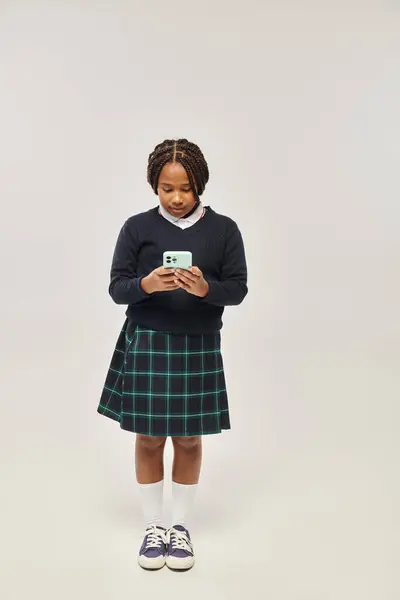 Preteen african american schoolgirl in uniform using smartphone on grey background, social media — Stock Photo