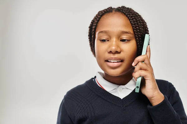 Preteen african american schoolgirl in uniform talking on smartphone on grey background, look down — Stock Photo