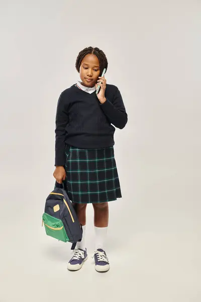 Preteen afrikanisch-amerikanische Schülerin in Uniform spricht auf Smartphone und hält Rucksack auf grau — Stockfoto