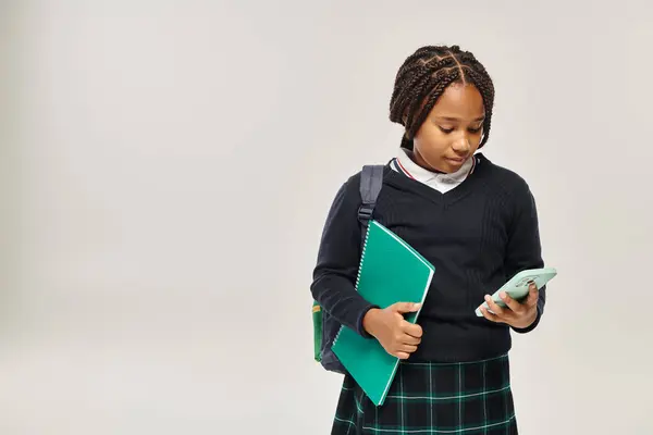 Pré-adolescente afro-américaine écolière en uniforme à l'aide d'un smartphone et tenant un carnet sur gris — Photo de stock