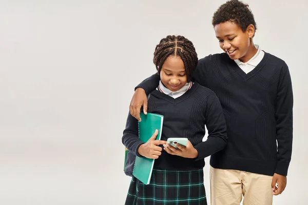 Щасливий афроамериканський школяр обіймає дівчину в уніформі зі смартфоном на сірому фоні — стокове фото