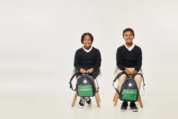 Cheerful african american schoolkids with backpacks sitting and looking at camera on grey backdrop — Stock Photo