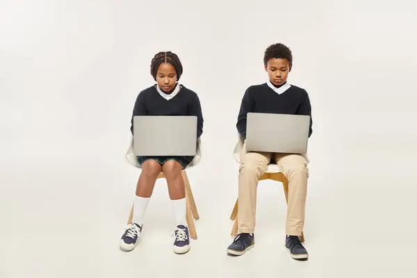 Concentrados alunos afro-americanos em uniforme usando laptops e sentados em pano de fundo cinza — Fotografia de Stock