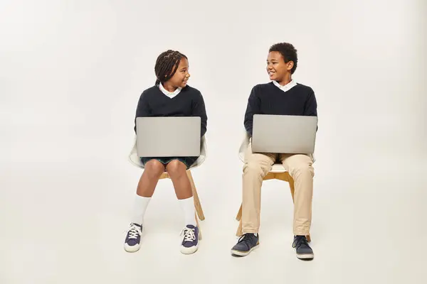 Allegro afroamericano scolari in uniforme utilizzando i computer portatili e seduto su sfondo grigio — Foto stock