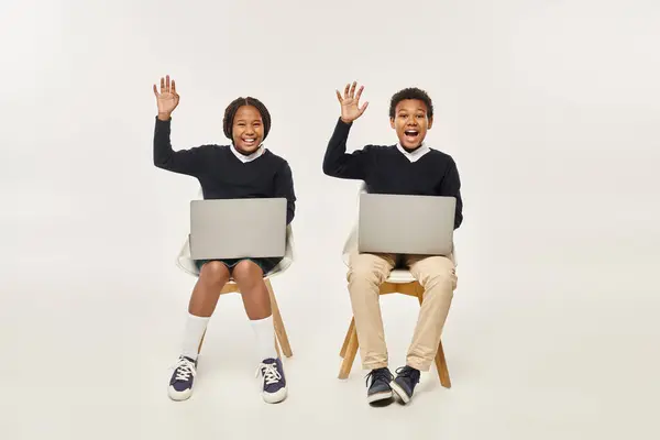 Aufgeregte afrikanisch-amerikanische Schulkinder in Uniform mit Laptops und winkenden Händen vor grauem Hintergrund — Stockfoto