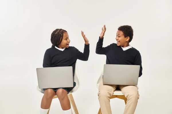 Fröhliche afrikanisch-amerikanische Schulkinder in Uniform mit Laptops und High Five vor grauem Hintergrund — Stockfoto