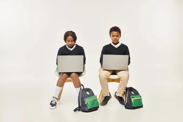 Зосереджені афроамериканські школярі в уніформі з використанням ноутбуків і сидячи на сірому фоні — Stock Photo