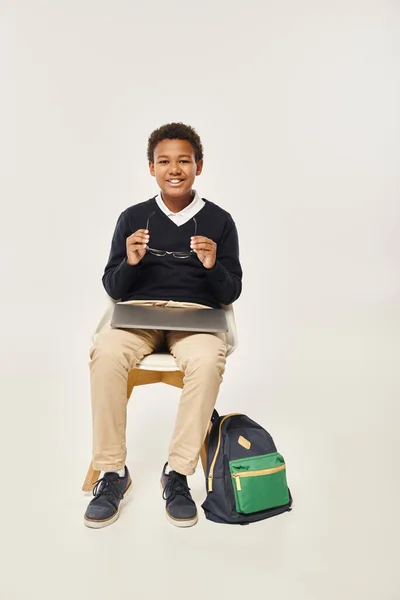 Happy african american schoolboy in uniform holding glasses and sitting with laptop on grey backdrop — Stock Photo