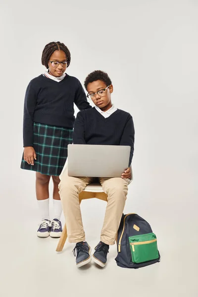 Cute african american girl in uniform standing near schoolboy using laptop on grey backdrop — Stock Photo