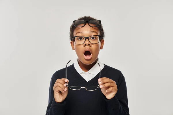 Shocked african american boy in sweater and eyewear holding glasses and looking at camera on grey — Stock Photo