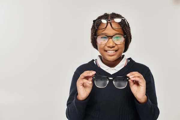Felice ragazza afroamericana in maglione e occhiali tenendo gli occhiali e guardando la fotocamera sul grigio — Foto stock
