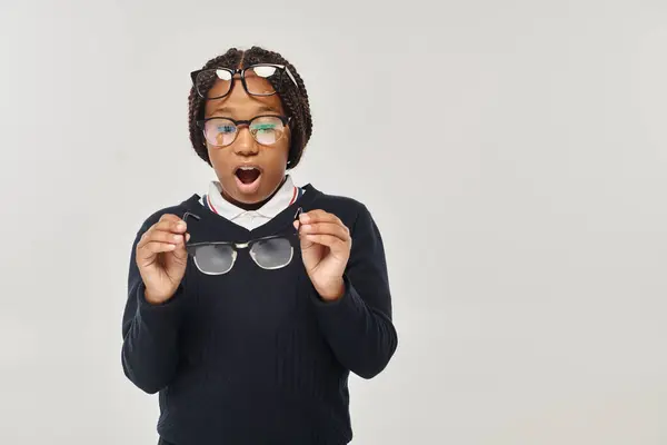 Shocked african american girl in sweater and eyewear holding glasses and looking at camera on grey — Stock Photo