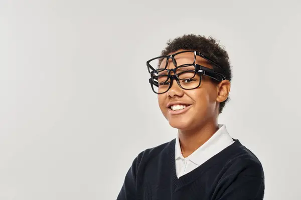 Optimistic african american boy in sweater and eyewear holding glasses and looking at camera on grey — Stock Photo