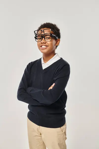 Cheerful african american boy in sweater and eyewear holding glasses and looking at camera on grey — Stock Photo