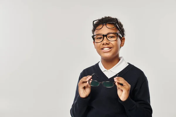 Happy african american schoolboy in eyewear holding glasses and looking at camera on grey backdrop — Stock Photo